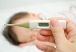 Mother holding a thermometer in the foreground with a sick child sleeping in the background