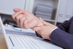 A man working at a computer, grabbing his right wrist in pain. Carpal tunnel syndrome occurs when the tissues in your wrist get swollen or inflamed and press on the median nerve, making your hand hurt.