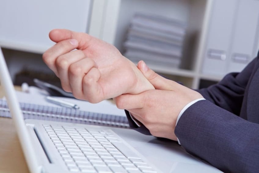 A man working at a computer, grabbing his right wrist in pain. Carpal tunnel syndrome occurs when the tissues in your wrist get swollen or inflamed and press on the median nerve, making your hand hurt.