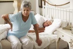 older woman sits on bed with her hand on her hip in pain