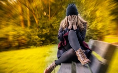 woman sitting on bench outside covering face with hands, surrounded by trees filled with anxiety