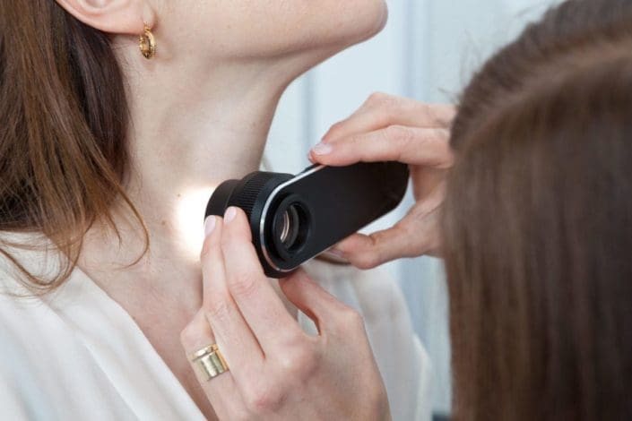 A doctor uses a lighted instrument to closely examine moles on a female patient’s neck