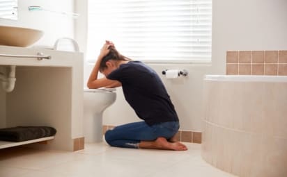 A woman kneeling on the bathroom floor while holding her head over the toilet as she vomits