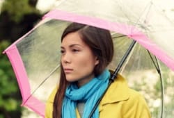 sad-looking woman in a coat and scarf stands under an umbrella in the rain
