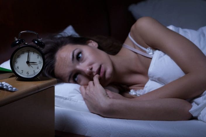 Woman lying in bed sleepless at night with clock and melatonin pills on her bedside table