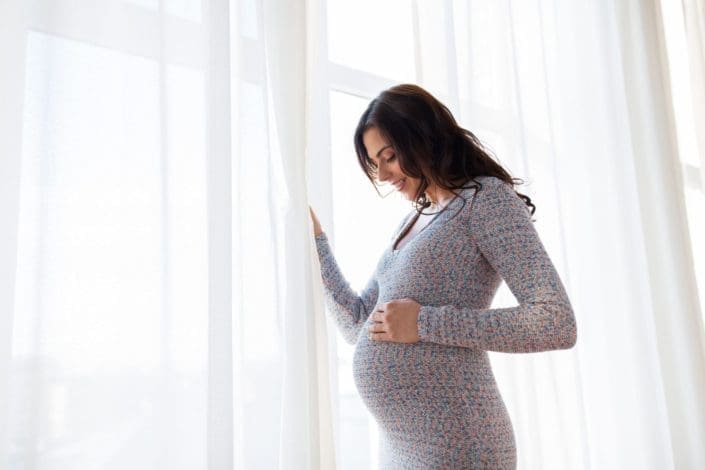 pregnant woman standing at window, smiling at belly