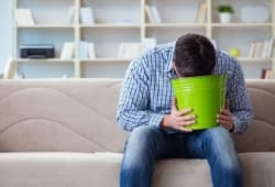 A man with food poisoning sits on the couch and vomits into a green bucket.