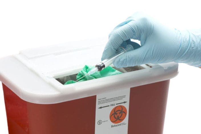 Gloved hand disposing of needle in hazardous waste container