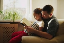 father and daughter sitting on couch reading a book together