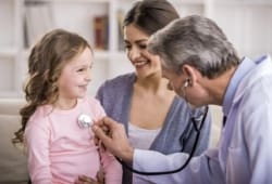 Family doctor using a stethoscope to examine a little girl in the arms of her mother.