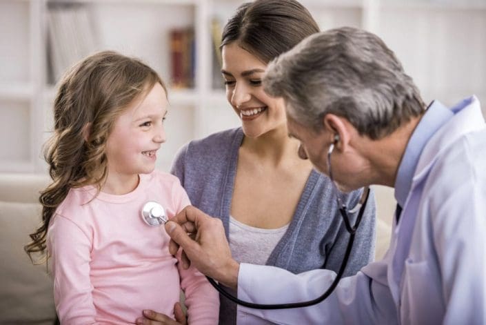 Family doctor using a stethoscope to examine a little girl in the arms of her mother.