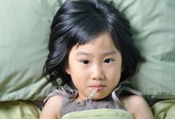 A little girl lies in bed with a thermometer in her mouth. Fifth disease is a mild viral infection that causes a bright red rash on the face. Other symptoms include fever, sore throat and headache.