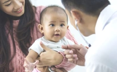 A mother holds baby being checked for bronchiolitis by a doctor with a stethoscope.