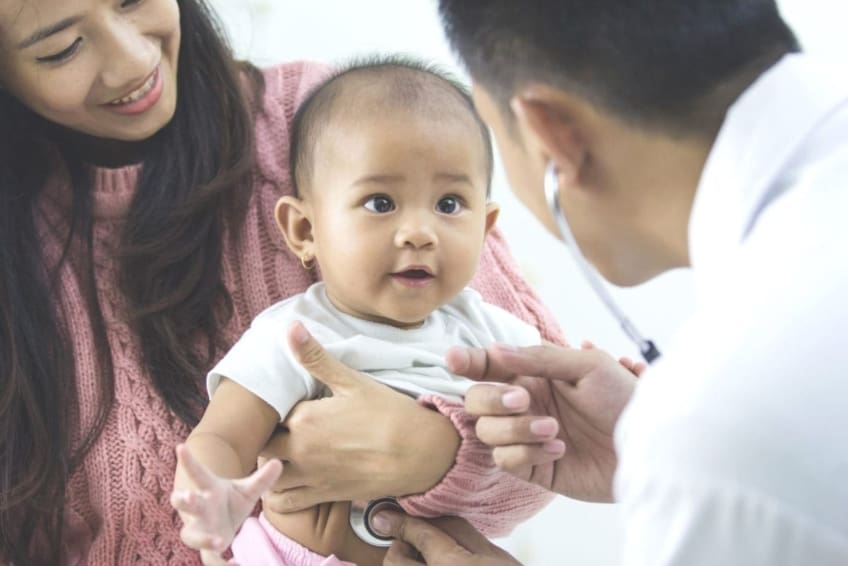 A mother holds baby being checked for bronchiolitis by a doctor with a stethoscope.