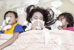 A mother and two young children lie in bed blowing their noses