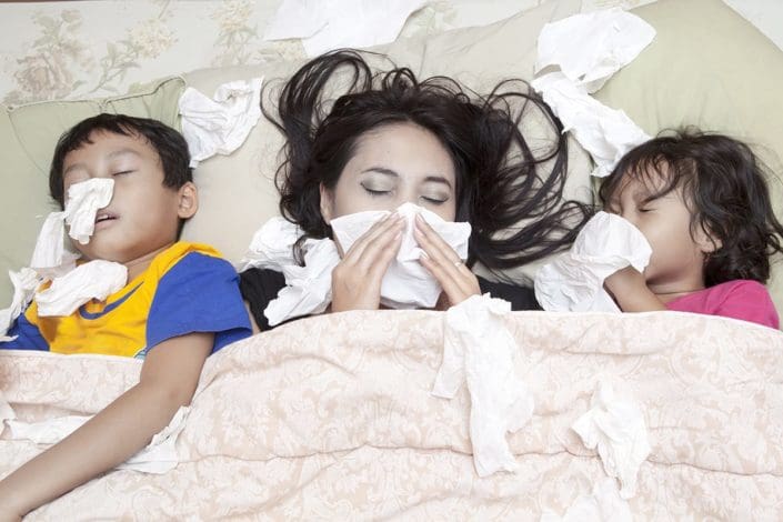 A mother and two young children lie in bed blowing their noses. Flu symptoms can sometimes be mistaken for symptoms of the common cold and vice versa. Both are contagious respiratory illnesses.