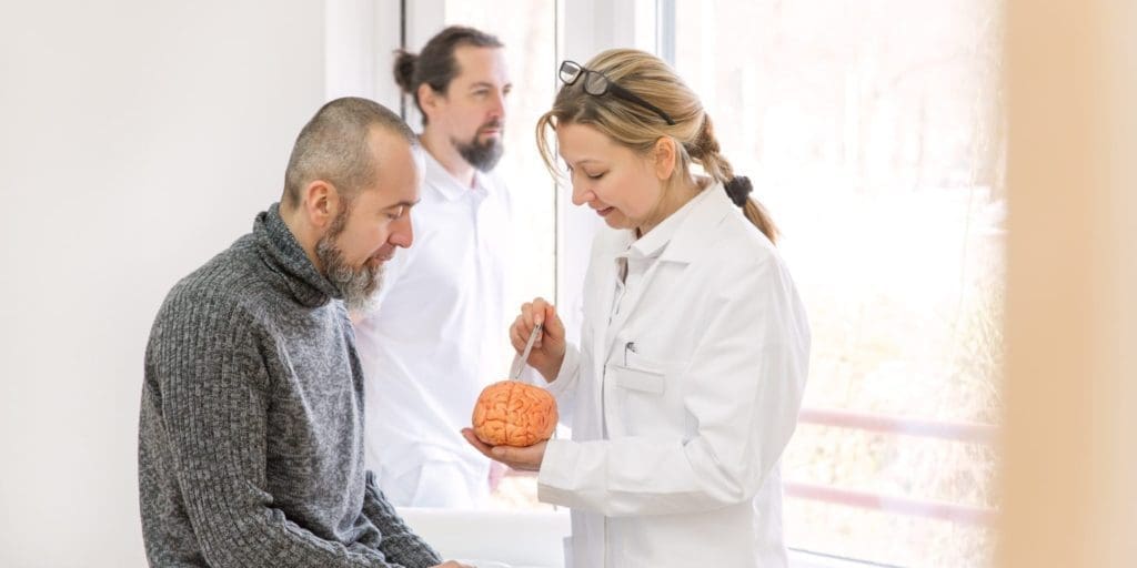 A female neurologist shows a male patient with multiple sclerosis symptoms something on a synthetic brain.