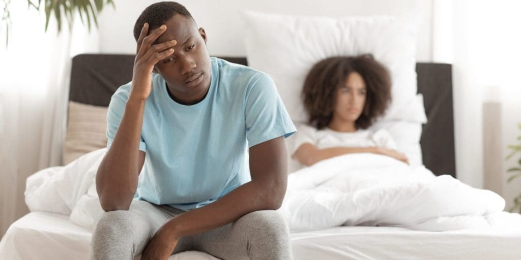 Male struggling with premature ejaculation sits on edge of bed with girlfriend in the background.