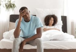 Male struggling with premature ejaculation sits on edge of bed with girlfriend in the background.