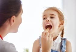 Doctor examining a child, checking her throat and tonsils. Common tonsillitis symptoms include redness and feeling like your throat or neck glands are swollen due to inflammation of the tonsils.