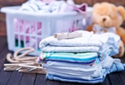 A laundry basket full of baby laundry and a stack of folded clothes sit on a wooden table.
