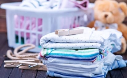 A laundry basket full of baby laundry and a stack of folded clothes sit on a wooden table.