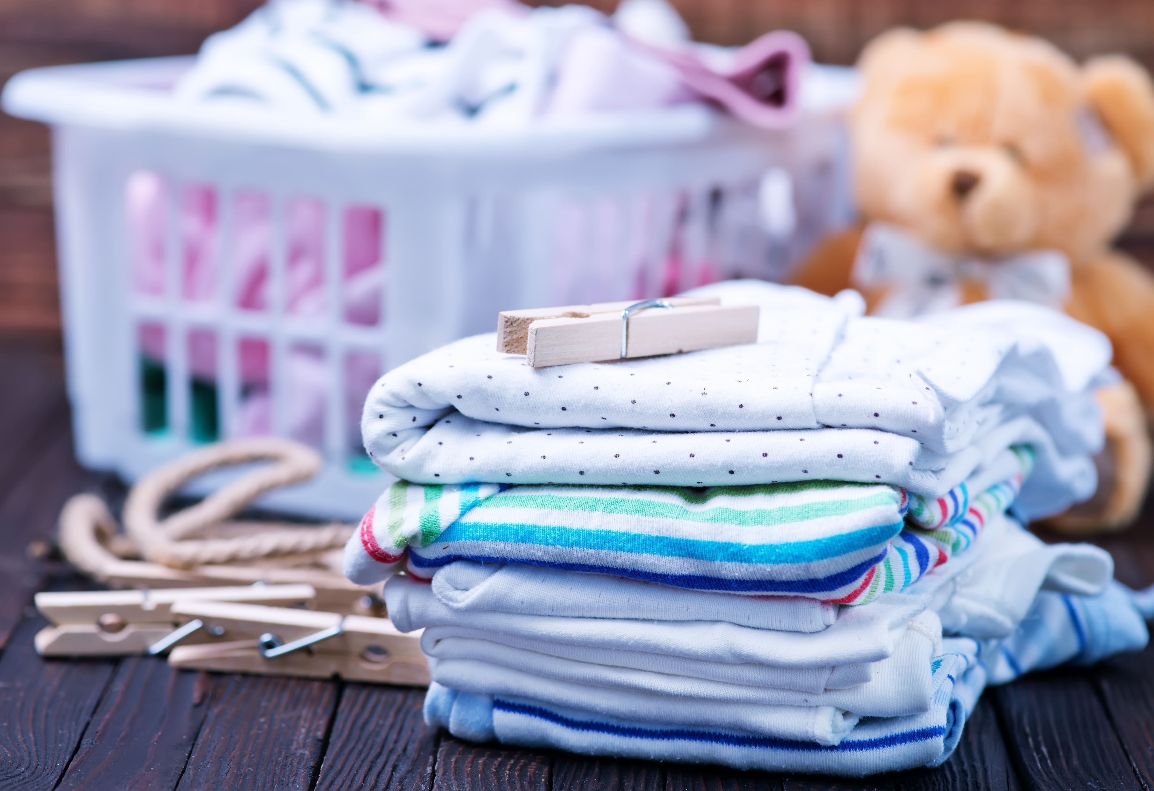 A laundry basket full of baby laundry and a stack of folded clothes sit on a wooden table.