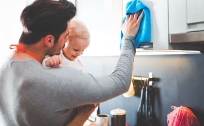 A young father cleans the kitchen while holding his daughter.