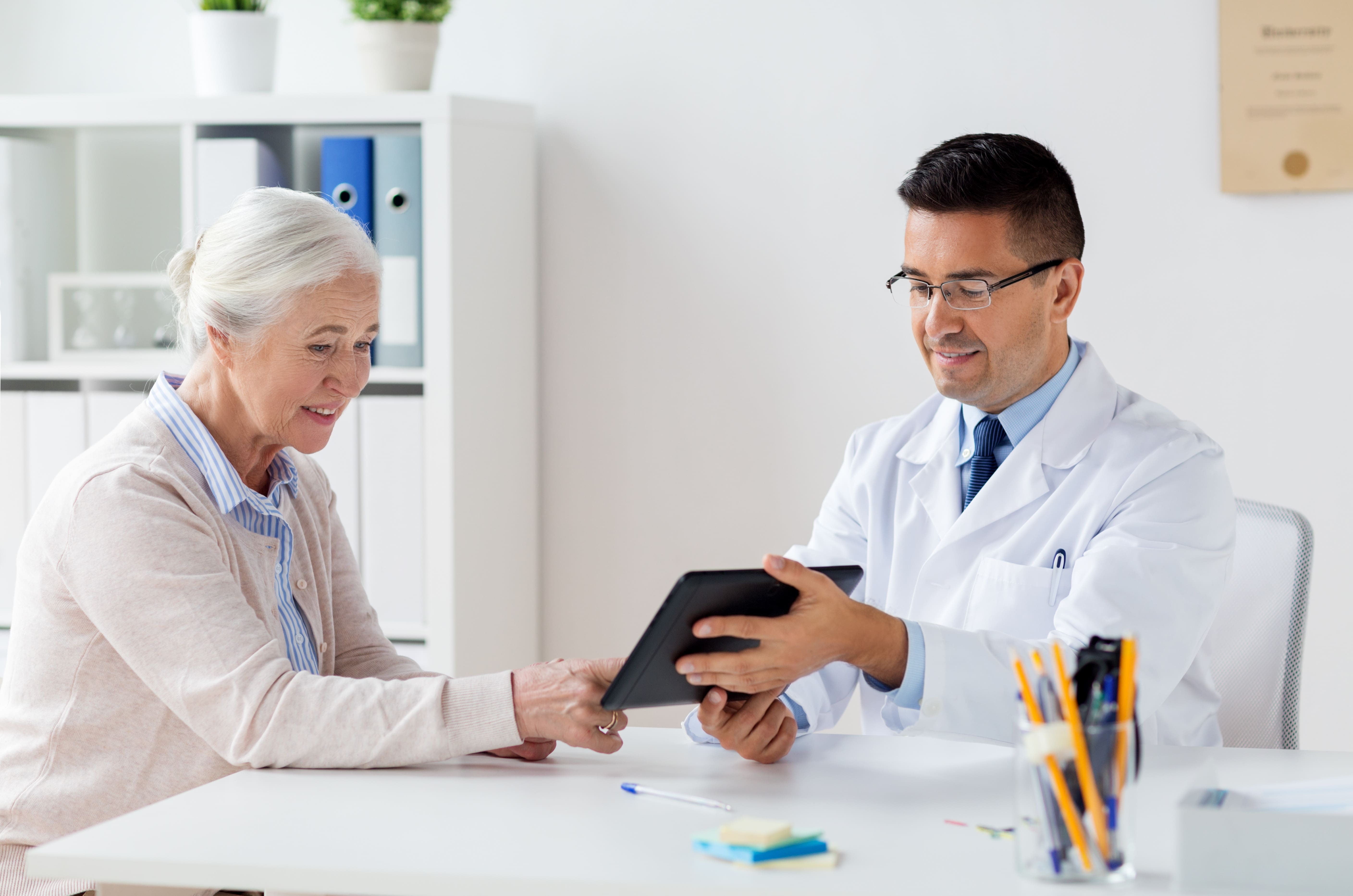 An elderly woman and doctor sit at a table and look at a tablet.