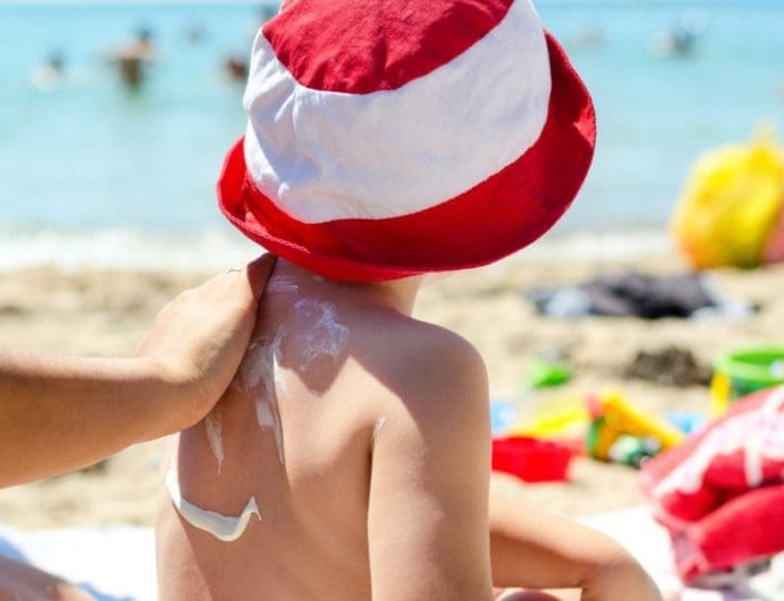 Young boy sitting on the beach having sunscreen applied to his back