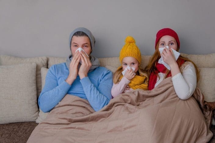 A family suffering from the flu holds tissues up to their noses while sitting on a couch.