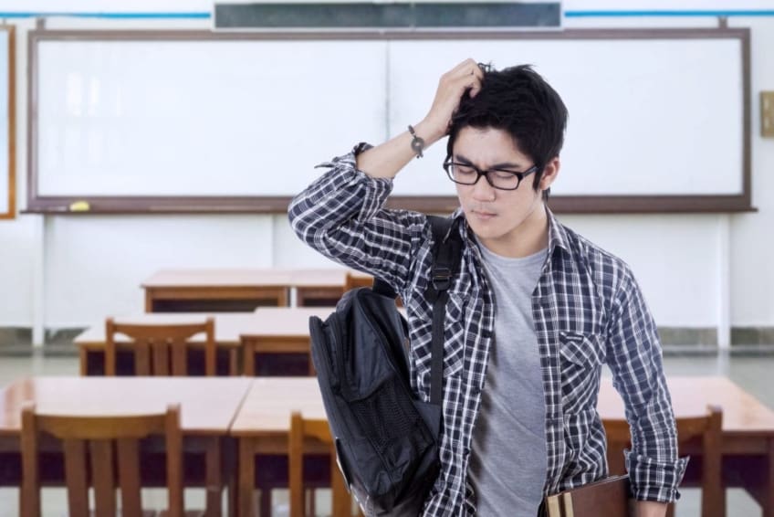 Teen boy standing in classroom wearing backpack, holding head in pain