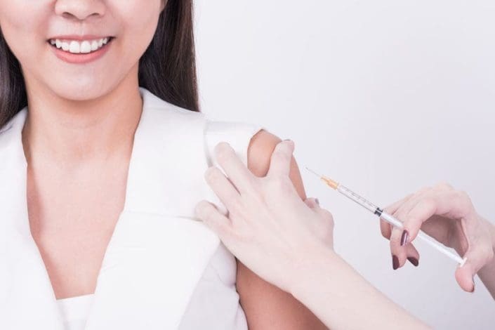 A woman with dark colored hair, wearing a white shirt receiving vaccination