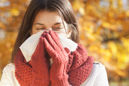 A woman with brown hair wearing orange gloves and standing outside by fall leaves blows her nose into a white tissue.
