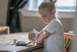 A fourteen year old Caucasian girl reads her blood sugar level by placing her cellphone next to her pump in her arm as she sits at a dining table.