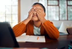 A girl sits at the kitchen table covering her eyes in a stressful way with her homework sitting in front of her.