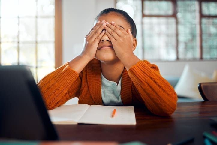 A girl sits at the kitchen table covering her eyes in a stressful way with her homework sitting in front of her.
