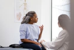 A woman, appearing worried, is seated on an exam table while talking with her doctor.
