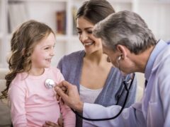 Family doctor using a stethoscope to examine a little girl in the arms of her mother.