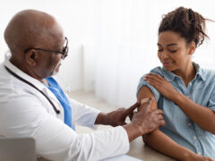 A doctor in a white lab coat with a stethoscope places a Band-Aid on the upper arm of a patient who just received a flu vaccine.