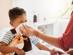 Flu, sick or cold child with parent sneezing, blowing and wiping runny nose while ill with covid virus, sinus and allergy symptoms in bed at home. Mother caring for stuffy and congested little son