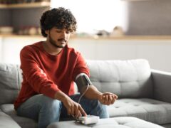Healthcare Concept. Young Indian Man Checking Blood Pressure With Upper Arm Monitor At Home, Millennial Eastern Guy Measuring Arterial Tension While Sitting On Couch In Living Room, Copy Space