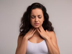 Sore throat, tonsilitis concept. Unhappy millennial brunette woman touching her neck, suffering from pain in throat, checking enlarged adenoids isolated on grey studio background, copy space
