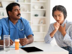 Sick Young Indian Woman Coughing During Meeting With Male Doctor In Clinic, Ill Female Patient Suffering Acute Bronchitis Or Pneumonia, Having Medical Check Up With Mature Therapist Man In Hospital