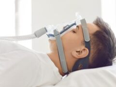 Close up side view of a male patient sleeping in a hospital bed with oxygen mask in white room. Young man recovering after sickness or getting obstructive sleep apnea therapy in hospital ward.