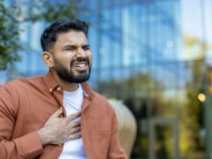 A man clutches his chest in pain, possibly experiencing a heart problem, in an outdoor setting. The background shows a blurred modern building.
