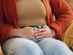 Hands, pain and person with stomach cramps, constipation and sick with irritable bowel syndrome on sofa. House, illness and woman with pelvic inflammatory disease, menstruation and touch of belly